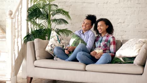 Two-mixed-race-curly-girl-friends-sitting-on-the-couch-and-watch-comedy-movie-and-eat-popcorn.-Funny-sisters-relaxing-together-at-home