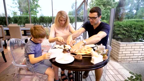 Padre-y-madre-se-sientan-en-la-mesa-con-su-hijo-y-da-rebanada-de-pizza-a-su-hijo-en-el-restaurante