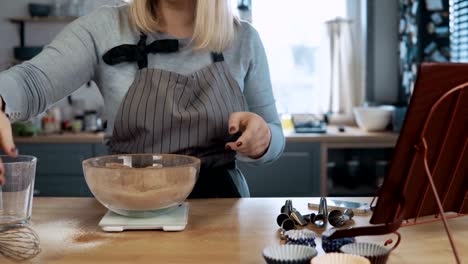 Junge-schöne-Frau-mischen-Zutaten-in-Schüssel-geben,-mit-Schneebesen.-Hübsche-Frau-Blick-auf-Rezept-vom-Kochen-Buch