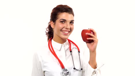 Young-friendly-female-nutritionist-showing-a-red-apple,-biting-it-and-smiling,-advising-healthy-diet.-Portrait-of-young-professional-with-stethoscope-and-lab-coat-isolated-on-white-background