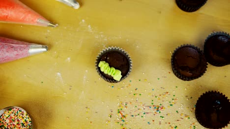 Vista-superior-de-las-manos-del-joven-decorando-los-cupcakes-de-chocolate-con-crema-de-colores.-Mujer-usando-la-Manga-pastelera