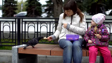 Mother-and-daughter-eat-sandwiches-and-feed-pigeons-on-a-cold,-cloudy-day.