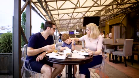 Image-of-a-happy-family-and-their-son-at-the-cafe.-Loving-couple-with-child-are-spending-time-together-in-pizzeria.-Mother,-father-and-son-relaxing-in-restaurant-eating-pizza-and-drinking-hot-tea