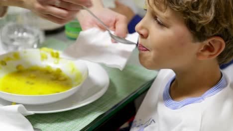 Mamá-alimenta-a-su-hijo-saludable-sopa-de-verduras-durante-la-cena-de-la-cena.-Niño-siendo-alimentado-con-comida-sana.-Escena-franca-e-informal-de-guapo-6-8-años-viejo-cena-comer-en-4K