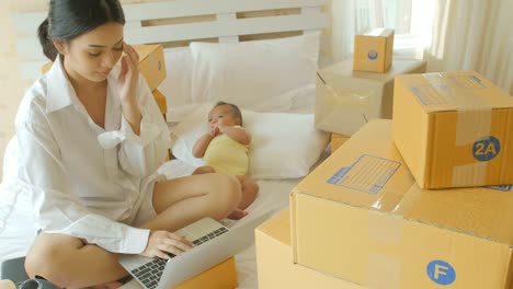 Young-asian-mother-and-her-newborn-child-checking-order-from-laptop-for-customer-and-online-delivery-for-ready-packing-in-bedroom.