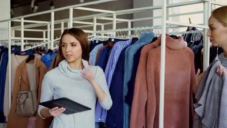 Young-businesswoman-is-using-tablet-while-checking-goods-in-her-clothing-store.-Assistant-is-coming-with-garment-and-asking-advice.-Business-owner-is-giving-orders.