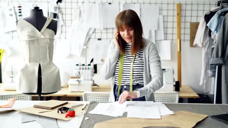 Young-attractive-dressmaker-is-discussing-clothing-designs-on-mobile-phone-and-checking-sketches-while-working-in-her-studio-between-dummy-and-rail-with-clothes.