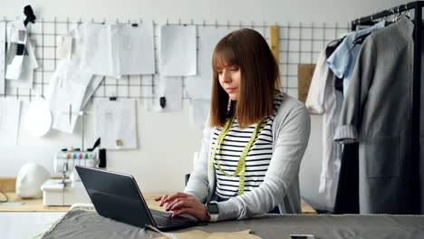 Young-self-employed-tailor-is-communicating-with-customers-using-laptop.-Good-view-of-light-modern-studio-in-background.-Successful-e-commerce-in-clothing-design-industry-concept.