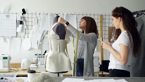 Young-woman-is-measuring-mannequin-parts-with-tape-measure-while-her-colleague-is-entering-this-information-in-smartphone.-Productive-cooperation-in-tailoring-business-concept.