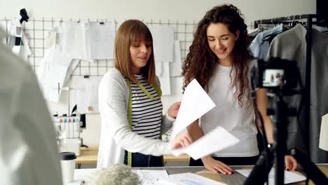 Junge-hübsche-Frauen-Modedesigner-Blogger-sind-mit-Kamera-über-neue-Kleidungsstück-Nähen-Videoaufnahme.-Mädchen-zeigen-Skizzen-und-Vorlagen-und-miteinander-reden.