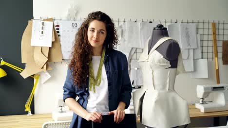 Portrait-of-beautiful-young-woman-tailor-standing-in-workplace-near-clothed-mannequin-and-looking-at-camera.-Women's-garments,-sketches-and-sewing-machines-are-visible.