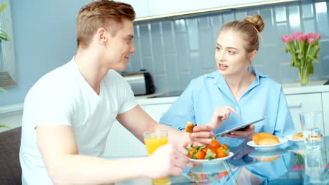 Pareja-joven-con-tableta-digital-desayunando-en-la-mesa-de-la-cocina.