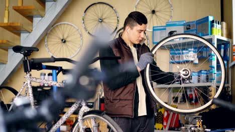 Mecánico-experimentado-está-girando-la-rueda-de-bicicleta-de-comprobación-de-trabajo-del-mecanismo-y-escuchando-música-mientras-trabajaba-en-el-pequeño-taller.-Concepto-de-mantenimiento-y-personas.