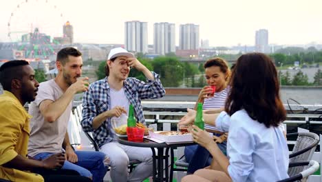 Multiracial-group-of-friends-is-clinking-glasses,-drinking-soft-drinks-and-eating-pizza-sitting-at-table-on-rooftop-celebrating-friends-reunion.-Food-and-party-concept.