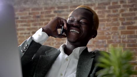 Young-blond-african-american-businessman-talking-on-phone-and-sitting-at-desk-with-laptop-in-modern-office,-laughing