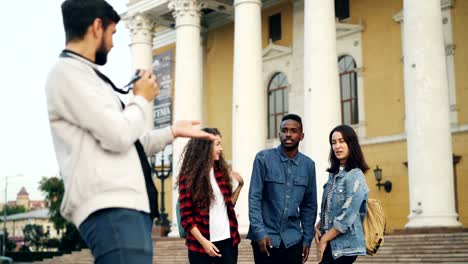 Fotógrafo-profesional-está-rodando-grupo-de-viajeros-felices-jóvenes-riendo-y-posando-para-la-cámara-delante-teatro-hermoso-con-mochilas.