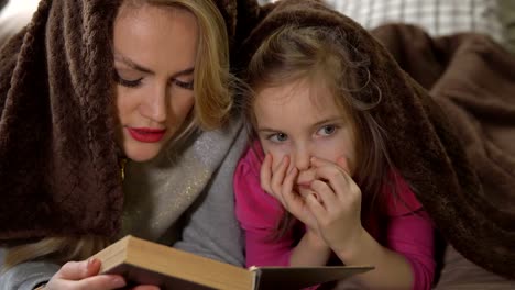 Madre-e-hija-se-encuentran-en-una-cama-bajo-una-manta-y-leer-un-libro.