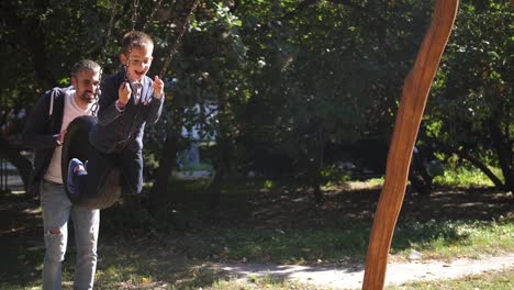 Glückliche-junge-Vater-und-seine-Kinder-auf-der-Schaukel-spielen-im-Park.-Glücklicher-lächelnder-Vater-rollt-seinen-Sohn-auf-einer-Schaukel-im-Park.-4-k.-Slow-motion