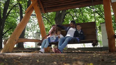 Glückliche-junge-Familie-Vater,-Mutter,-Sohn-und-Töchterchen-reitet-auf-einer-Schaukel-im-Park.-Lachen-und-Lächeln.-4-k.-Slow-Motion