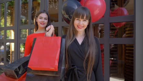 shopping,-smiling-young-woman-showing-many-of-paper-bags-with-purchases-on-background-of-pretty-girlfriend-with-balloons