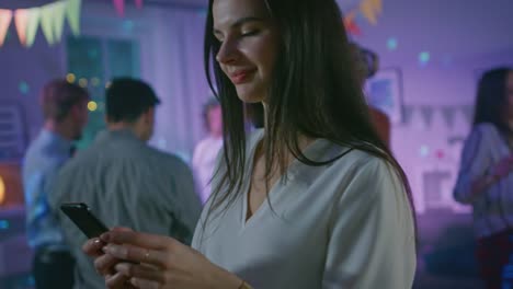 At-the-Wild-House-Party:-Beautiful-Young-Girl-Uses-Smartphone,-Browses-Through-Internet-and-Social-Network.-In-the-Background-Crowd-of-Young-People-Dancing-Off-and-Having-Fun.