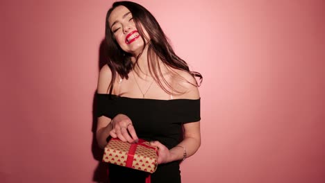 Brunette-happy-woman-open-her-present-with-confetti-in-pink-background-wear-black-dress