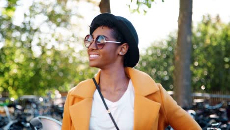 Moda-joven-mujer-negro-permanente-al-aire-libre-llevando-gafas-de-sol,-una-capa-amarilla-y-un-sombrero-negro-mirando-a-la-cámara-y-riendo,-cierre-para-arriba,-centrarse-en-primer-plano