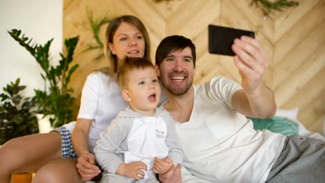 Padres-sonriendo-con-el-bebé-tomando-foto-de-la-familia-selfie-en-la-cama-en-casa