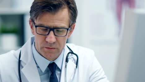 Close-up-of-a-Male-Senior-Doctor-Working-at-His-Desk-on-a-Personal-Computer.-Reflection-of-the-Screen-Seen-in-His-Glasses.