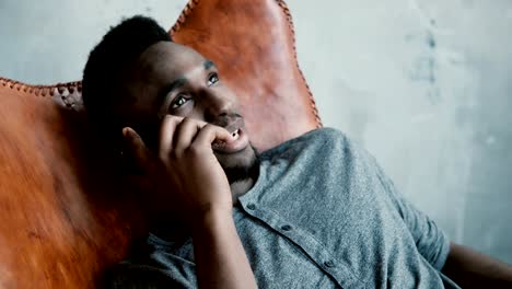 Portrait-of-young-African-male-with-a-beard-sitting-in-chair,-using-his-Smartphone.-Man-talks-on-phone-and-smiles