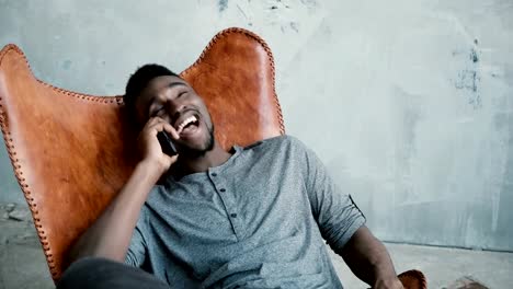 Portrait-of-African-man-sitting-in-the-chair-and-talking-on-the-Smartphone.-Male-is-laughing-at-joke,-having-great-time