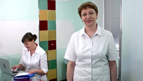 Woman-doctor-smiling-and-looking-to-the-camera-while-her-colleague-is-working-on-laptop