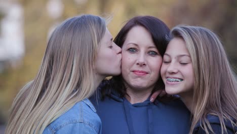 Atractiva-madre-mirando-a-cámara-con-dos-hijas-muy-jóvenes-dándole-un-beso-en-la-mejilla