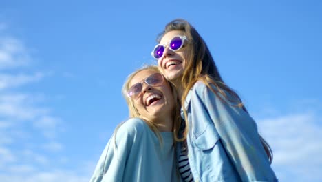 Dos-hermosas-chicas-felizes-llevando-gafas-de-sol-abrazan,-ríen-y-tienen-un-montón-de-diversión.-Cielo-azul-de-fondo.