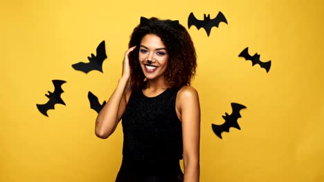 Pretty-afro-american-lady-with-cat-make-up-and-ears-laughing-and-flirting-to-camera-isolated-over-yellow-wall-with-bats