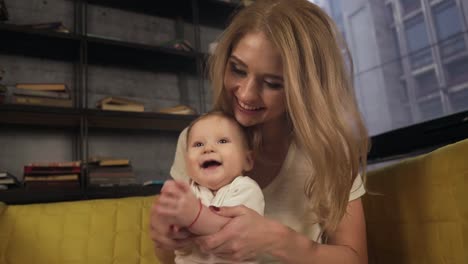 Lovely-mother-is-sitting-on-yellow-sofa,-holding-her-baby-boy-and-playing-with-him-on-hands.-The-baby-is-smiling-and-laughing.-Front-view,-portrait