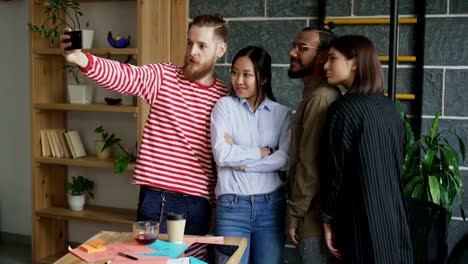 Happy-multi-ethnic-team-taking-selfie-in-modern-loft-office.-Hipster-man-holding-smartphone-take-self-portrait-with-his-colleagues