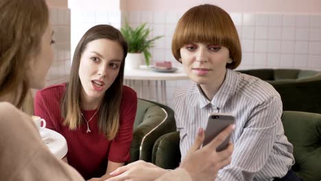 Three-young-caucasian-girls-are-sitting-in-a-cafe,-friends,-company,-gossips,-dialogue,-discussion.-Girlfriends-in-the-cafe-concept.-Using-a-smartphone,-yes-by-shaking-head-60-fps