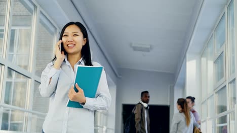 Retrato-de-joven-hermosa-estudiante-de-etnia-asiática-hablando-por-teléfono-sonriendo-positivamente-pie-en-corredor-de-colegio-con-sus-amigos-detrás-de