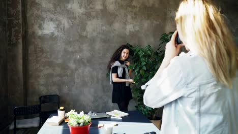 Chica-atractiva-es-posando-con-planta-grande-mientras-colega-photogrpahing-le-en-cámara-digital-en-la-oficina-moderna-lof.-Las-mujeres-están-divirtiéndose-y-riendo-durante-la-pausa-café