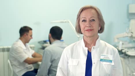 Senior-femenino-médico-mirando-a-cámara-mientras-hombre-doctor-hablando-con-el-paciente-en-el-fondo