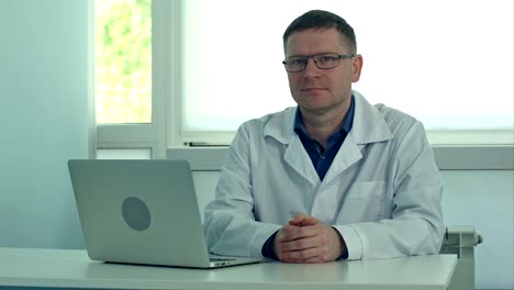 Mature-male-doctor-sitting-at-desk-with-laptop-and-looking-at-camera-in-his-clinic-office