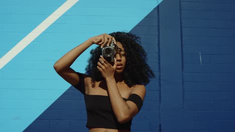 Beautiful-young-woman-shooting-photos-with-film-camera-against-colorful-blue-wall