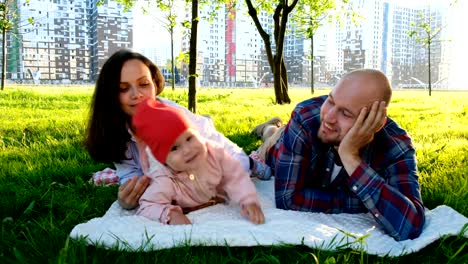 Joven-madre-besa-a-una-hija-pequeña-en-el-parque-en-verano,-el-padre-se-ríe-y-relojes.-Familia-feliz-con-el-bebé-descansando-en-el-parque-de-verano-al-atardecer