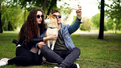 Handsome-young-man-is-taking-selfie-with-his-pretty-wife-and-cute-dog,-all-wearing-sunglasses.-Guy-is-holding-smartphone-taking-pictures-and-posing.