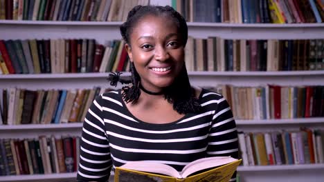 Libro-de-lectura-de-joven-hermosa-mujer-Afro-Americana-en-biblioteca,-Sonriente,-feliz,-Fondo-de-estanterías