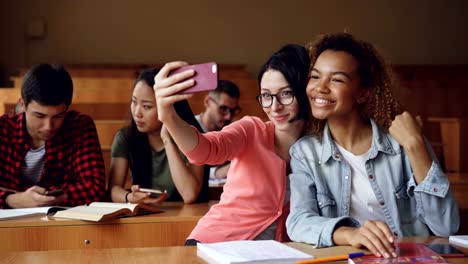 Fröhliche-Mädchen-Studenten-nehmen-Selfie-mit-Smartphone-sitzen-an-Tischen-am-College,-Frauen-posieren-Hand-Gesten-und-umarmt.-Freunde-und-Technologiekonzept.