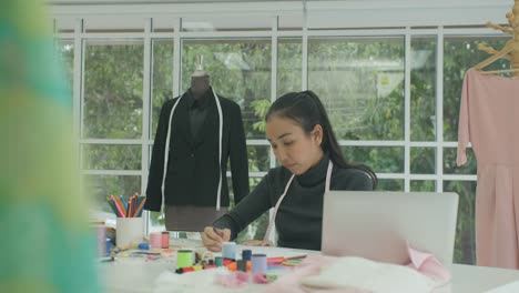 Diseñador-de-moda-de-mujer-asiática-DS-MS-hermoso-sentado-en-su-escritorio-mirando-bocetos-en-un-ordenador-portátil-en-estudio,-tomando-notas