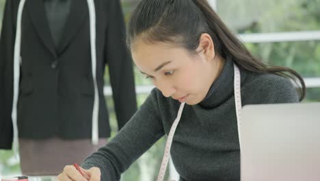 Beautiful-asian-woman-fashion-designer-sitting-at-her-desk-looking-at-sketches-on-a-laptop-in-modern-studio,-Taking-notes