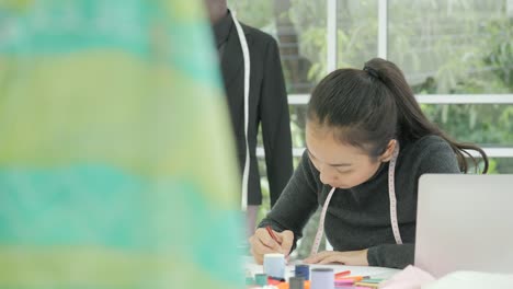 DS-MS-Beautiful-asian-woman-fashion-designer-sitting-at-her-desk-looking-at-sketches-on-a-laptop-in-modern-studio,-Taking-notes